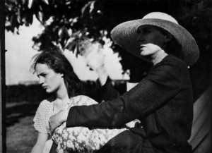Black and white photo of a girl and a woman sitting outside in bright sun, the woman in a dark dress and broad hat